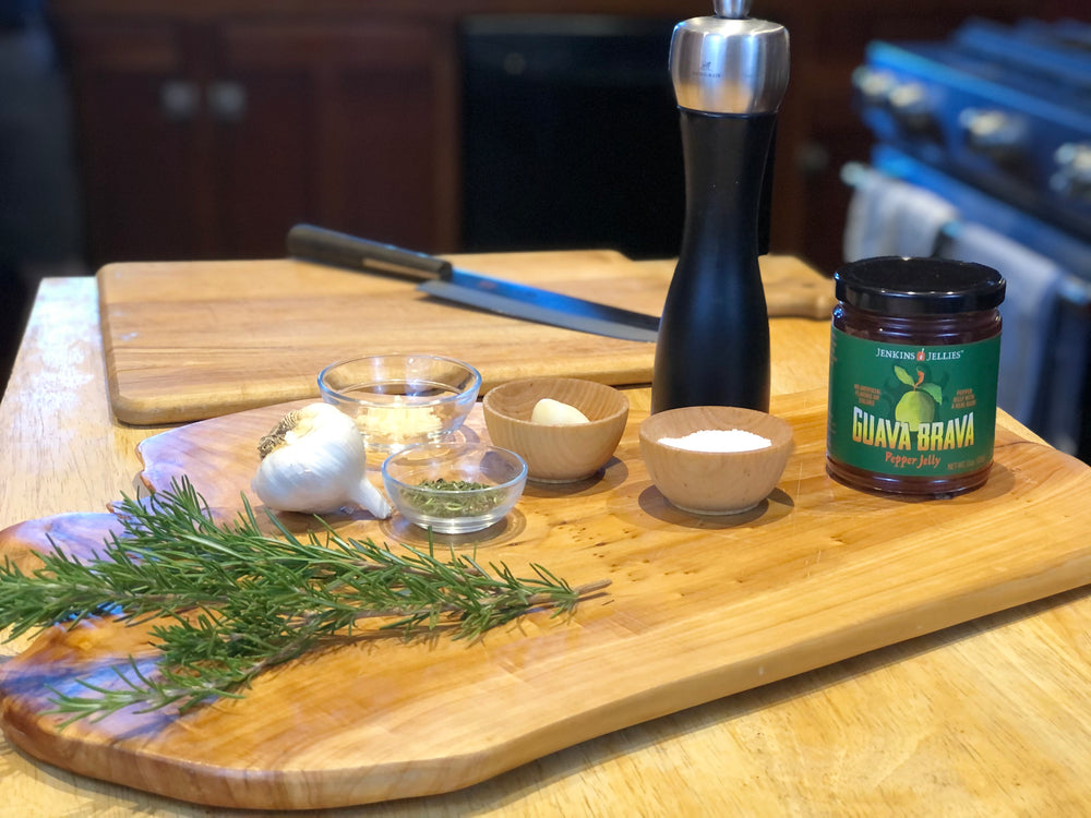 
                  
                    Load image into Gallery viewer, 11 oz jar of Guava Brava sweet and spicy hot chili jelly sitting on a wooden cutting board with garlic, fresh rosemary, salt and pepper. Jenkins Jellies Guava hot jam is used as a pantry staple in this kitchen. Food prep for the home chef.
                  
                