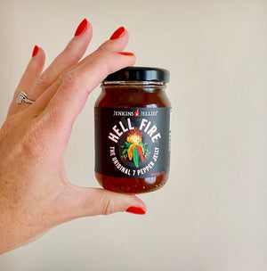 
                  
                    Load image into Gallery viewer, 5 oz. jar of Jenkins Jellies Hell Fire pepper jelly held in one hand to show the size. Wedding ring and manicured nails with red nailpolish compliment the painted hot chillis on the product label.
                  
                