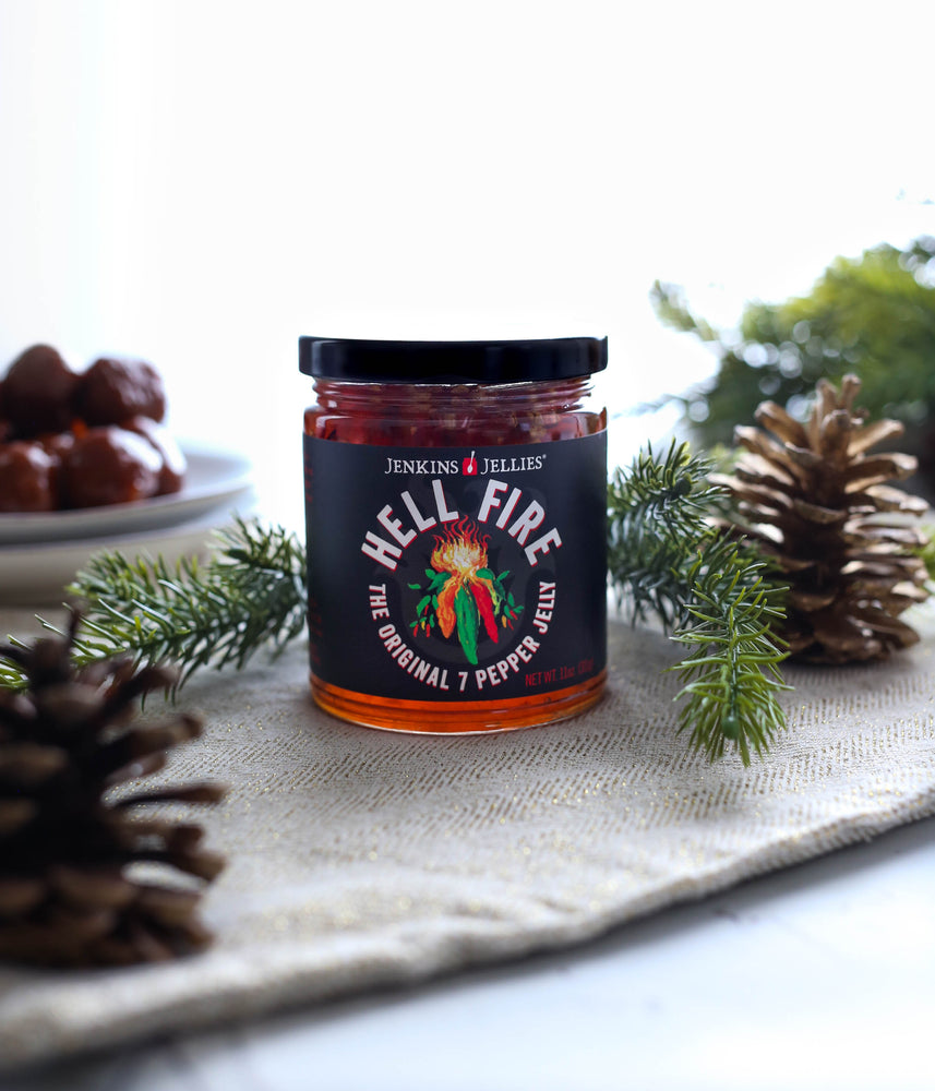 
                  
                    Load image into Gallery viewer, A full 11 oz jar of Jenkins Jellies Original 7 pepper jelly. This perfectly balanced sweet and savory hot pepper jam is sitting on a holiday decorated table. With evergreen cuttings and pinecones with party meatballs in the background. Perfect pepper jam for your holiday parties. Make it extraordinary.
                  
                