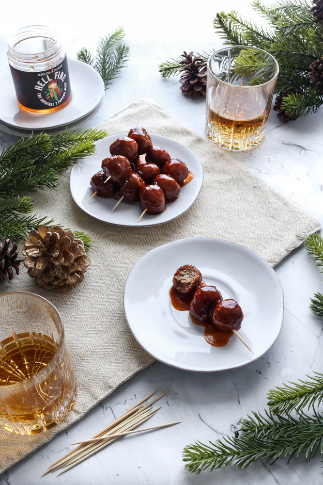 Hell Fire pepper jam is shown here on festive holiday table with evergreen shrub, pinecones and a whiskey cocktail. The jar of hot pepper jelly is empty because it was used in a sauce that cooked holiday meatballs for the party. Guests enjoyed party sized meatballs drenched in mouthwatering sweeet and spicy bbq sauce with hot pepper jam. 