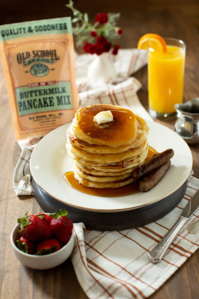 Pictured is a tall stack of delicious Old School Brand pancakes with syrup and butter dripping down the sides. There is also a bowl of strawberries and a tall glass of fresh squeezed orange juice.