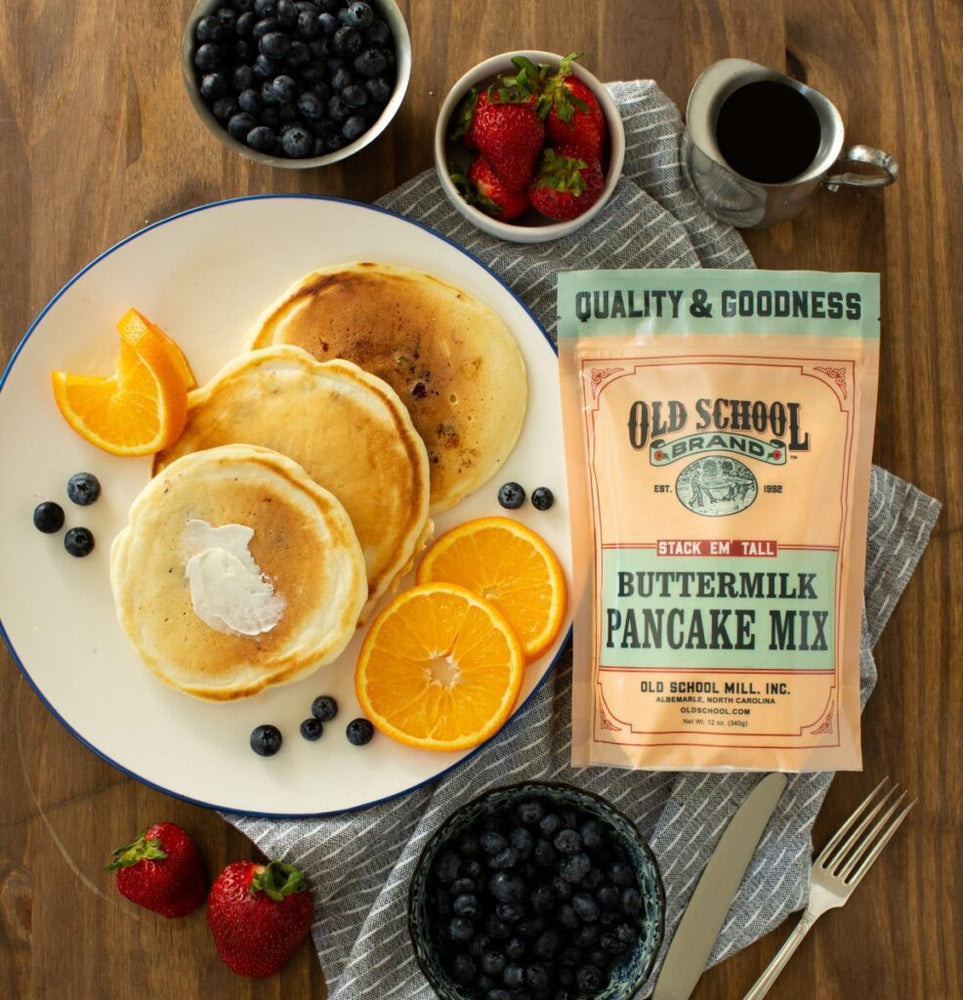 Bird's eye view of Old School Buttermilk pancake mix. Also shown is cooked pancakes with fresh fruit slices of oranges and bowls of blueberries and strawberries.