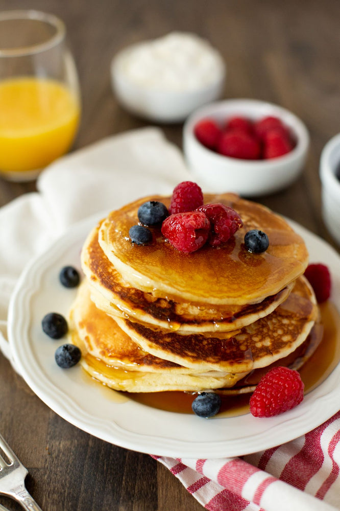 
                  
                    Load image into Gallery viewer, Stack of Old School brand pancackes with syrup and fresh raspberries and blueberries. Bowl of strawberries and glass of orange juice.
                  
                